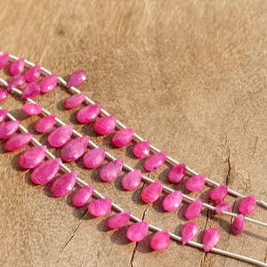 May include: A strand of pink gemstone beads in a teardrop shape. The beads are strung on a silver wire and are arranged in three rows.