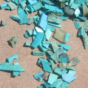 May include: A collection of turquoise blue gemstone chips scattered on a light brown surface. The chips are various shapes and sizes, some are rectangular, some are triangular, and some are irregular.