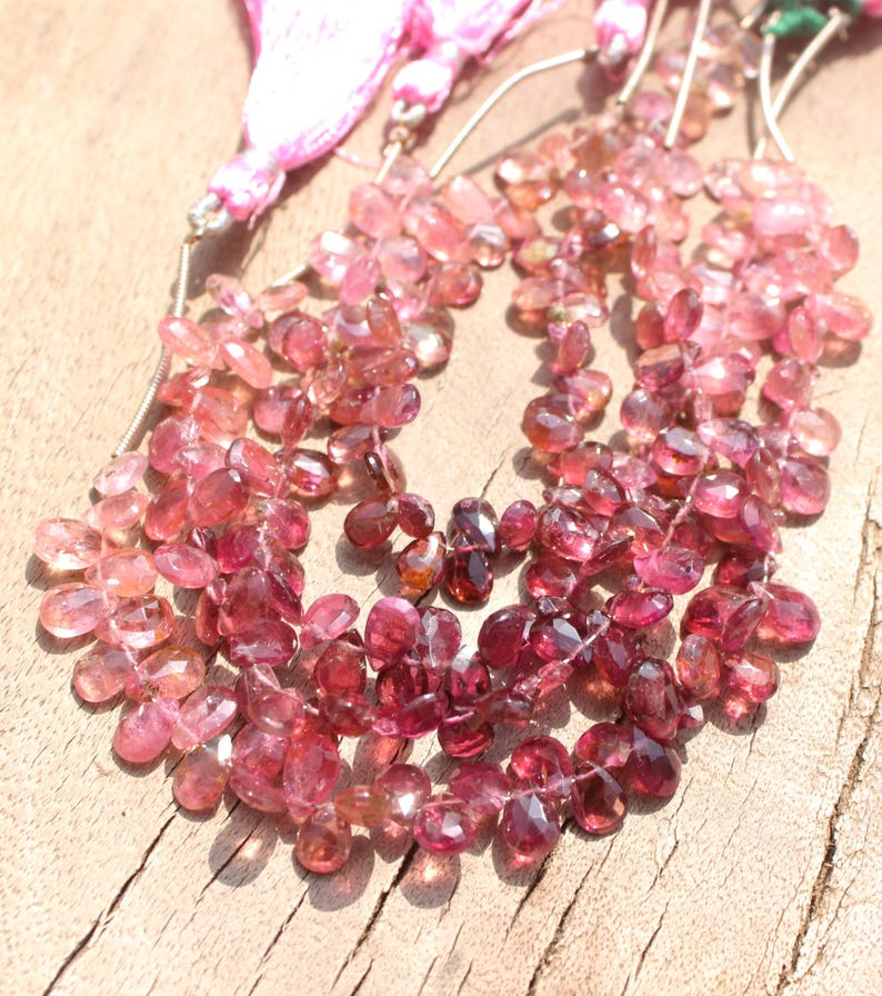 May include: A strand of pink and red gemstone beads, with a faceted, teardrop shape. The beads are strung on a silver wire and are ready to be used for jewelry making.