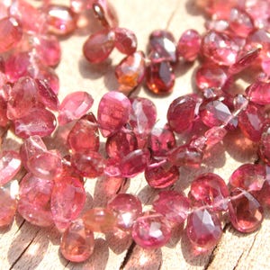 May include: A strand of pink tourmaline gemstone beads. The beads are faceted and have a teardrop shape. The beads are strung on a clear thread.