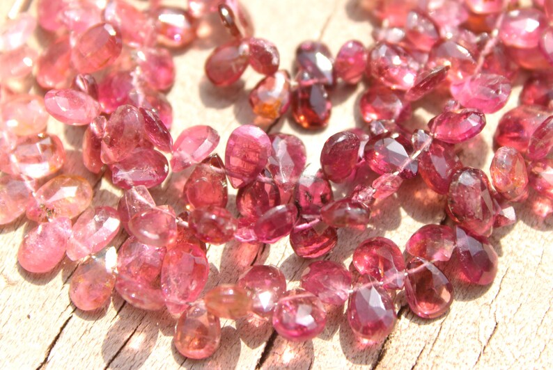 May include: A strand of pink tourmaline gemstone beads. The beads are faceted and have a teardrop shape. The beads are strung on a clear thread.