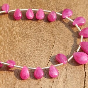 K&ouml;nnte beinhalten: Eine Reihe von rosafarbenen Rubin-Edelsteinkugeln in Tropfenform. Die Perlen sind auf einem Silberdraht aufgef&auml;delt und in einem Kreis angeordnet.