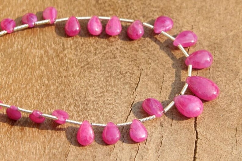 K&ouml;nnte beinhalten: Eine Reihe von rosafarbenen Rubin-Edelsteinkugeln in Tropfenform. Die Perlen sind auf einem Silberdraht aufgef&auml;delt und in einem Kreis angeordnet.
