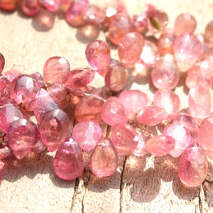 May include: A strand of pink gemstone beads, each bead is faceted and has a teardrop shape. The beads are strung on a clear thread and are arranged in a loose, flowing pattern.