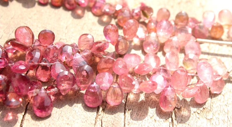 May include: A strand of pink gemstone beads, each bead is faceted and has a teardrop shape. The beads are strung on a clear thread and are arranged in a loose, flowing pattern.