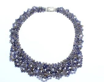 May include: A necklace composed of small, round, blue-gray beads. The beads are strung together in multiple strands, creating a layered effect. A silver-colored clasp secures the necklace. The jewelry is set against a white background.