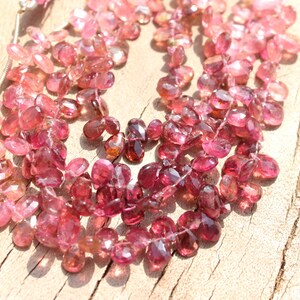 May include: A strand of pink and red gemstone beads, with a faceted, teardrop shape. The beads are strung on a clear thread and are arranged in a loose, flowing pattern.
