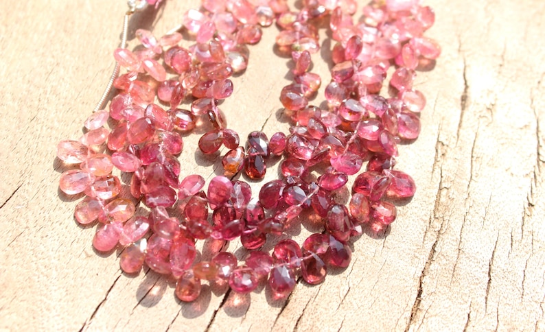 May include: A strand of pink and red gemstone beads, with a faceted, teardrop shape. The beads are strung on a clear thread and are arranged in a loose, flowing pattern.