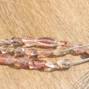 Puede incluir: Dos hilos de cuentas transparentes de forma ovalada con inclusiones rojas. Las cuentas están ensartadas en alambre de plata, con una borla colorida en el extremo. Las cuentas se muestran sobre una superficie de madera.