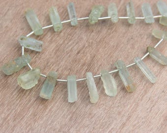 May include: A strand of raw, light green aquamarine gemstone beads. The beads are irregularly shaped, with a rough, natural texture. They are strung on a thin, silver-colored wire, and are displayed on a light brown wooden surface.