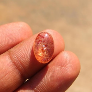 以下が含まれることがあります： オレンジと赤の混合色を持つ、楕円形のサンストーンの宝石。 研磨された石は、キラキラと光る半透明の外観で、目に見えるインクルージョンがあります。 宝石は手で持たれており、その自然な美しさと色のバリエーションを示しています。