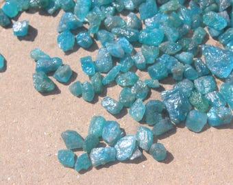 May include: A collection of small, irregularly shaped, translucent blue-green gemstones scattered on a light brown surface. The stones vary in size and have a rough, crystalline texture, catching the light and creating highlights.