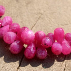 Op de afbeelding: Een ketting met een streng gefacetteerde, felroze edelstenen. De edelstenen zijn een mix van ronde en druppelvormige vormen, geregen op een zilveren ketting. De ketting wordt gepresenteerd op een houten oppervlak.