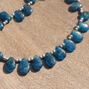 Puede incluir: Collar con piedras preciosas azules en forma de lágrima, ensartadas en una cadena plateada. Las piedras pulidas varían en tamaño y se muestran sobre una superficie de madera.