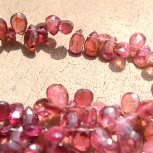 May include: A strand of pink and red gemstone beads, each bead is faceted and has a teardrop shape. The beads are strung on a thin wire and are arranged in a loose, flowing pattern.