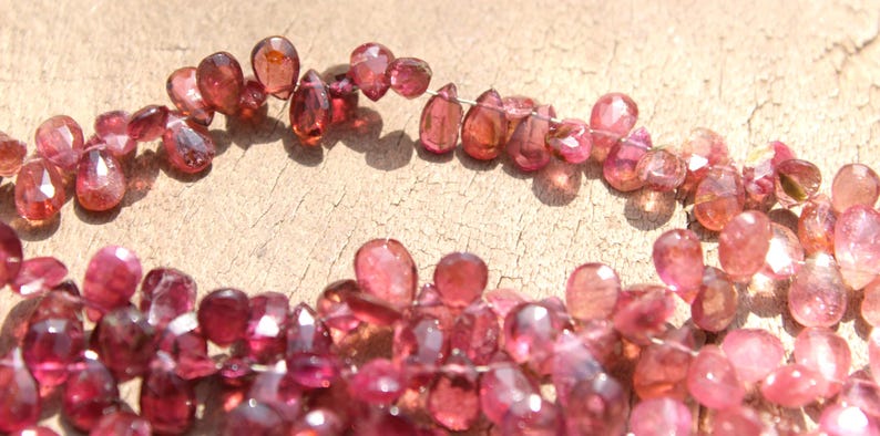 May include: A strand of pink and red gemstone beads, each bead is faceted and has a teardrop shape. The beads are strung on a thin wire and are arranged in a loose, flowing pattern.