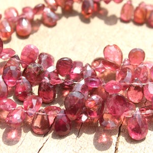 May include: A strand of pink gemstone beads, each bead is faceted and shaped like a teardrop. The beads are strung on a clear thread and are arranged in a loose, flowing pattern.