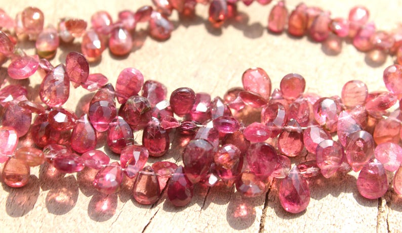 May include: A strand of pink gemstone beads, each bead is faceted and shaped like a teardrop. The beads are strung on a clear thread and are arranged in a loose, flowing pattern.