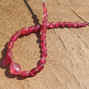 以下が含まれることがあります： 楕円形で半透明なピンク色の宝石のネックレス。ビーズは銀色のワイヤーに通され、ループを形成しています。宝石は、木目が粗い明るい茶色の木製の表面に展示されています。