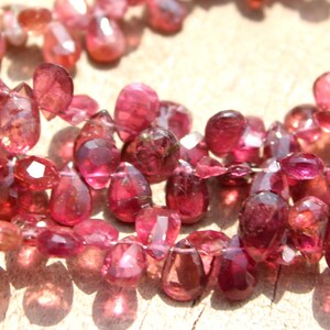 May include: A strand of pink and red gemstone beads, each bead is faceted and shaped like a teardrop. The beads are strung on a clear thread.