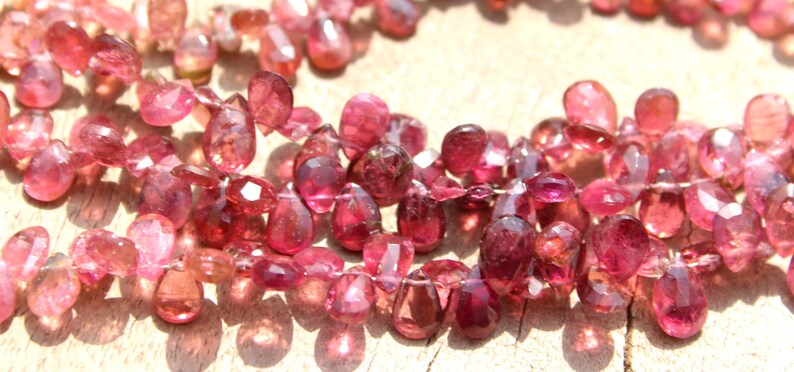 May include: A strand of pink and red gemstone beads, each bead is faceted and shaped like a teardrop. The beads are strung on a clear thread.