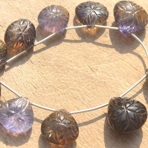 May include: A strand of carved gemstone beads in shades of purple and brown. The beads are heart-shaped and have a floral design. The beads are strung on a silver wire.
