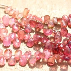 May include: A strand of pink and red gemstone beads. The beads are faceted and have a teardrop shape. The beads are strung on a silver wire.