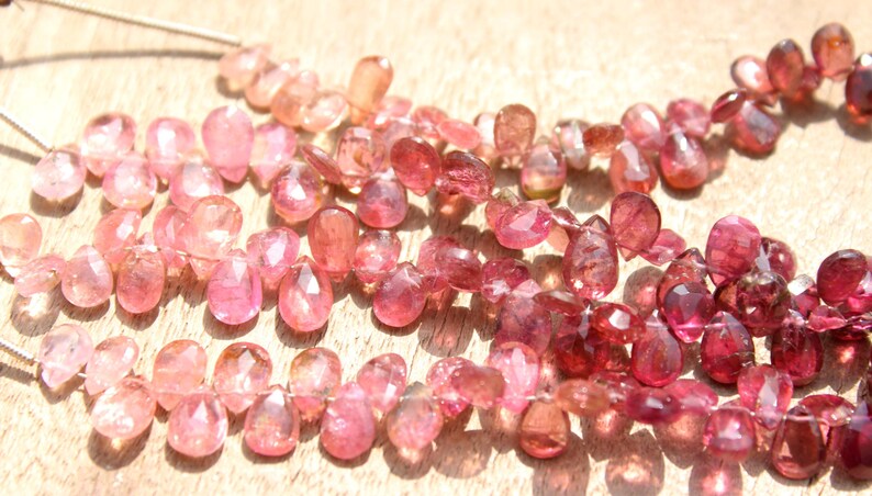 May include: A strand of pink and red gemstone beads. The beads are faceted and have a teardrop shape. The beads are strung on a silver wire.