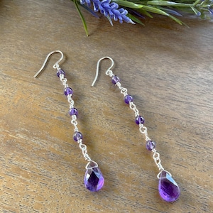 May include: A pair of dangle earrings featuring amethyst beads and teardrop-shaped pendants. The earrings have a silver wire design and are displayed on a wooden surface with purple flowers in the background. The earrings are approximately 3 inches long.