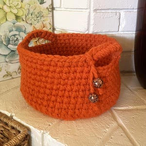 May include: An orange crochet basket with handles. The basket has a rounded shape and is adorned with two decorative, patterned buttons on the side. The basket is sitting on a white surface.