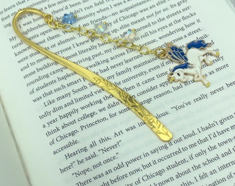 May include: A gold-colored metal bookmark with a decorative floral design and a charm featuring a white and blue pegasus. The bookmark has a chain with blue and clear beads. The bookmark is resting on an open book.