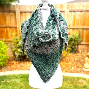 May include: A triangular crochet shawl featuring a textured stitch pattern in alternating bands of dark forest green and heather gray yarn. The garment is draped over a white mannequin, displaying a repeating cluster design throughout the fabric.