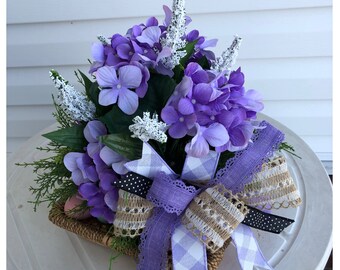 May include: A purple and white floral arrangement in a woven basket with a large purple and white bow. The arrangement features hydrangeas and sprigs of white flowers.