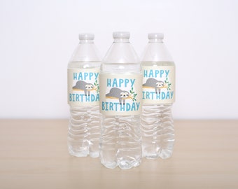May include: Three clear plastic water bottles with labels that say "Happy Birthday" and feature a cartoon sloth hanging from a branch. The labels are beige with blue text and green leaves.