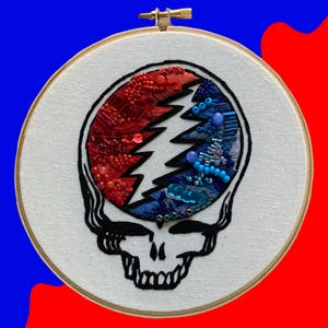 May include: Embroidered Grateful Dead Steal Your Face skull design with red, blue, and white beads on a white canvas in a wooden embroidery hoop.