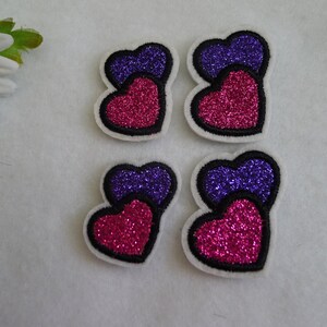 May include: Four glittery heart-shaped patches, each with a smaller purple heart on top of a larger pink heart. The hearts are outlined in black and have a white backing.