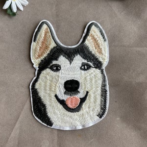 May include: Embroidered patch featuring a smiling husky dog face. The patch has a white background with black, brown, and cream-colored details. The dog's tongue is pink, and the patch is outlined in black.