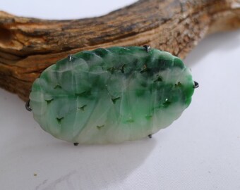 May include: An oval-shaped brooch featuring a carved green and white stone with a leaf-like pattern. The stone is secured by small metal prongs along its edges, creating a decorative piece with natural color variations and intricate surface textures.