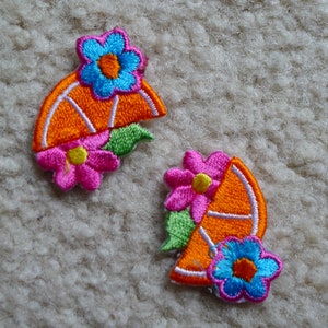 May include: Two embroidered iron-on patches featuring orange half-circles with white lines, pink and blue flowers, and green leaves.