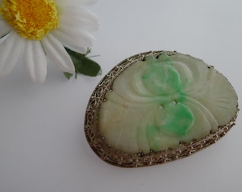 May include: Oval-shaped jade brooch with a carved floral design and a filigree metal border. The jade features shades of white and green. A white daisy with a yellow center is in the background.