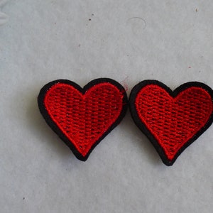 May include: Two red heart-shaped embroidered patches with black borders. The hearts have a textured appearance, and the patches are likely for decorative purposes, such as embellishing clothing or accessories. The background is a light, neutral color.