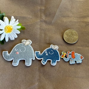 May include: Three embroidered elephant patches in shades of blue and gray, with white stitching and a small bow on their heads. One patch has a butterfly on its trunk. A white daisy and a coin are also in the image.