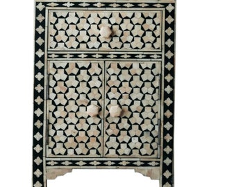 May include: A small, black and white bone inlay cabinet with a geometric pattern. The cabinet has one drawer and two doors with bone inlay knobs.
