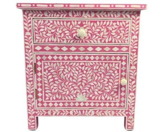 May include: A pink wooden nightstand with a floral bone inlay design. The nightstand has one drawer and one door. The drawer and door have bone inlay floral designs. The nightstand has a white bone inlay trim around the top and bottom.