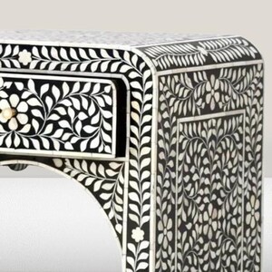May include: A black and white side table with a floral design made of bone inlay. The table has one drawer and a curved top.