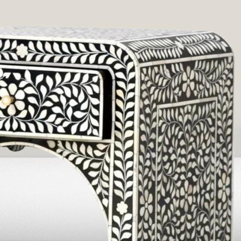 May include: A black and white side table with a floral design made of bone inlay. The table has one drawer and a curved top.
