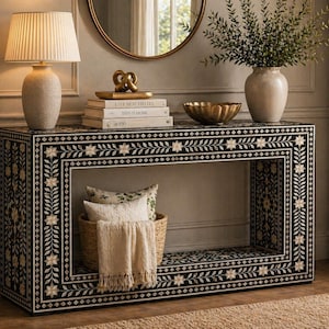 May include: A black and white floral patterned console table with a lamp, books, and decorative items on top. A woven basket with pillows and a blanket is placed below. A vase with greenery is also visible.