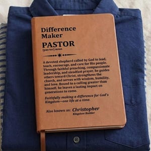 May include: A tan leather-bound journal featuring the printed definition of a pastor as a difference maker, resting on a folded navy blue button-down shirt. The text includes the name Christopher and the phrase Kingdom Builder in black ink.