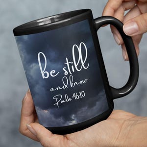 May include: Black ceramic mug with the words "be still and know Psalm 46:10" in white script. The mug features a dark blue cloudy sky design. The mug is held by a person.