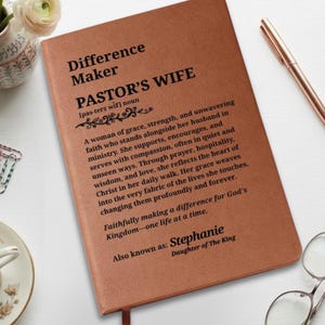 May include: A tan leather-bound journal featuring black printed text that reads Difference Maker PASTOR'S WIFE, followed by a definition, and the personalised name Stephanie with the phrase Also known as: Daughter of The King at the bottom.